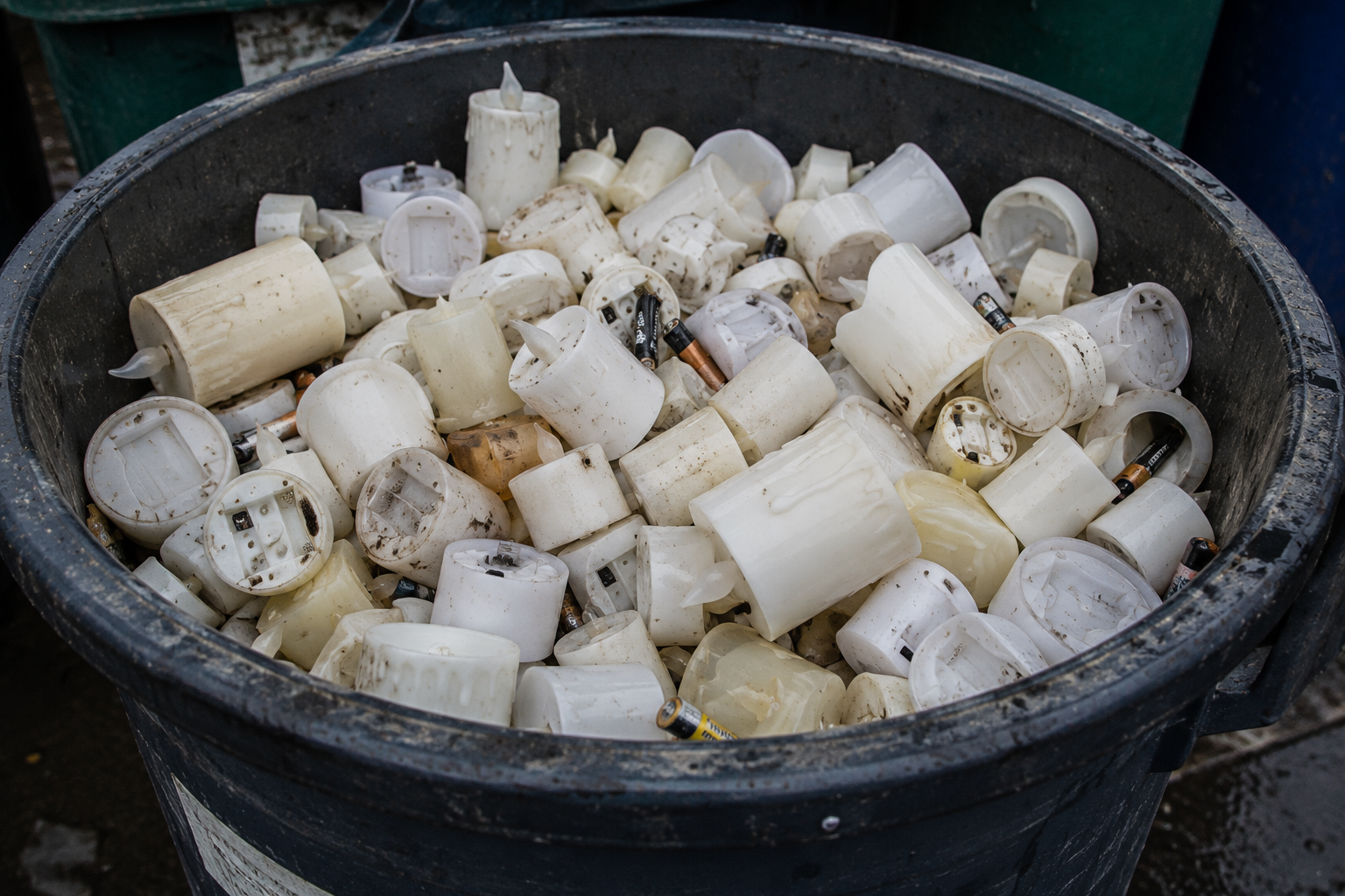 A large outdoor trash bin overflowing with discarded LED candles, plastic housings and used batteries. The image illustrates the amount of electronic waste generated by disposable battery-powered memorial candles.